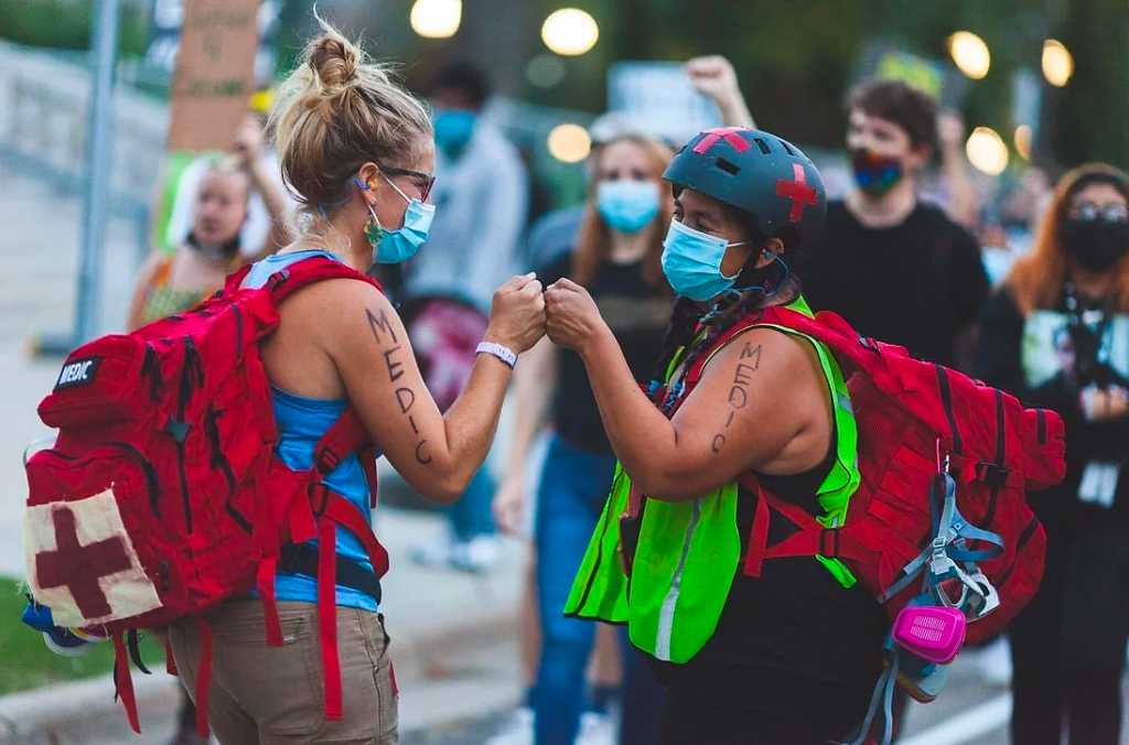 Two medics from North Star Medical wearing large, red backpacks and blue N95 masks, bump fists on a busy street with marchers in the background.