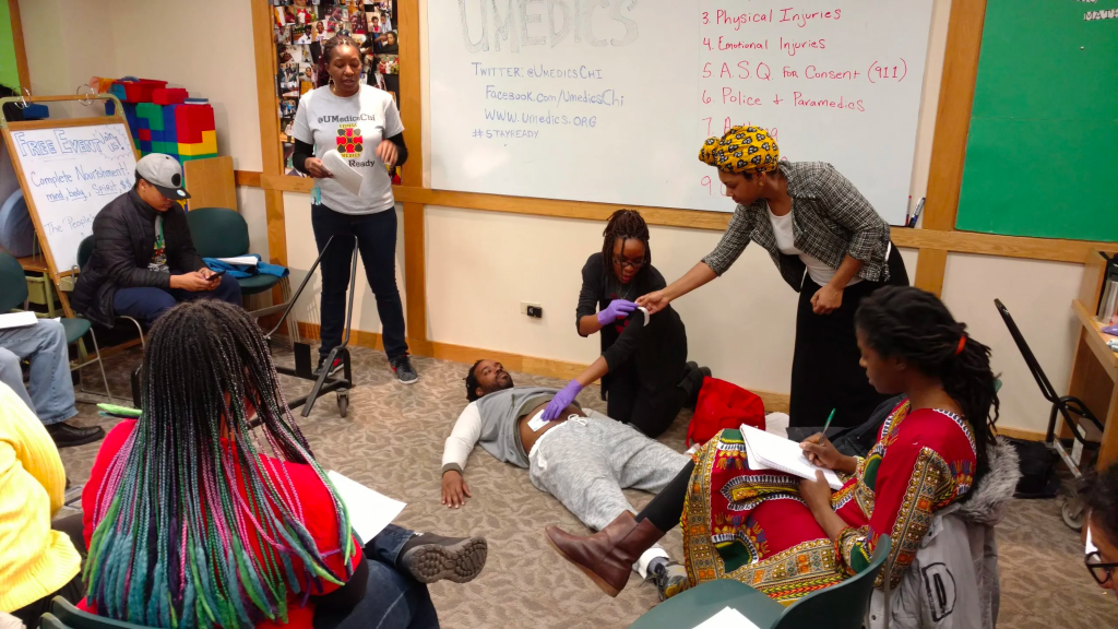 Image Description: Participants in a gunshot wound training take notes as two members of the Black-led Ujimaa Medics demonstrate the proper placement of wound dressings on a third facilitator who is lying on the ground. They are in a classroom accented with bright colors, and behind them is a whiteboard with a partially-obscured list that reads: (Three) Physical Injuries; (Four) Emotional Injuries; (Five) A.S.Q. For Consent (911); (Six) Police + Paramedics. And, to the left: UMEDICS; TWITTER: @UMEDICSCHI; Facebook.com/UmedicsChi; www.umedics.org; #StayReady