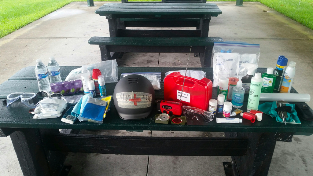 Medic supplies including a marked helmet, radio, wound kits, hand sanitizer, goggles, and bottled water are laid out on a dark green picnic table.