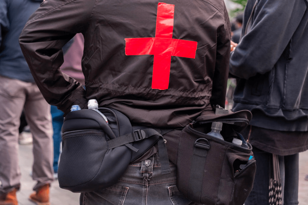 A medic wearing all black with several bags of water strapped to their waist stands with their back to the camera; a red cross in duct tape is displayed prominently on their jacket.