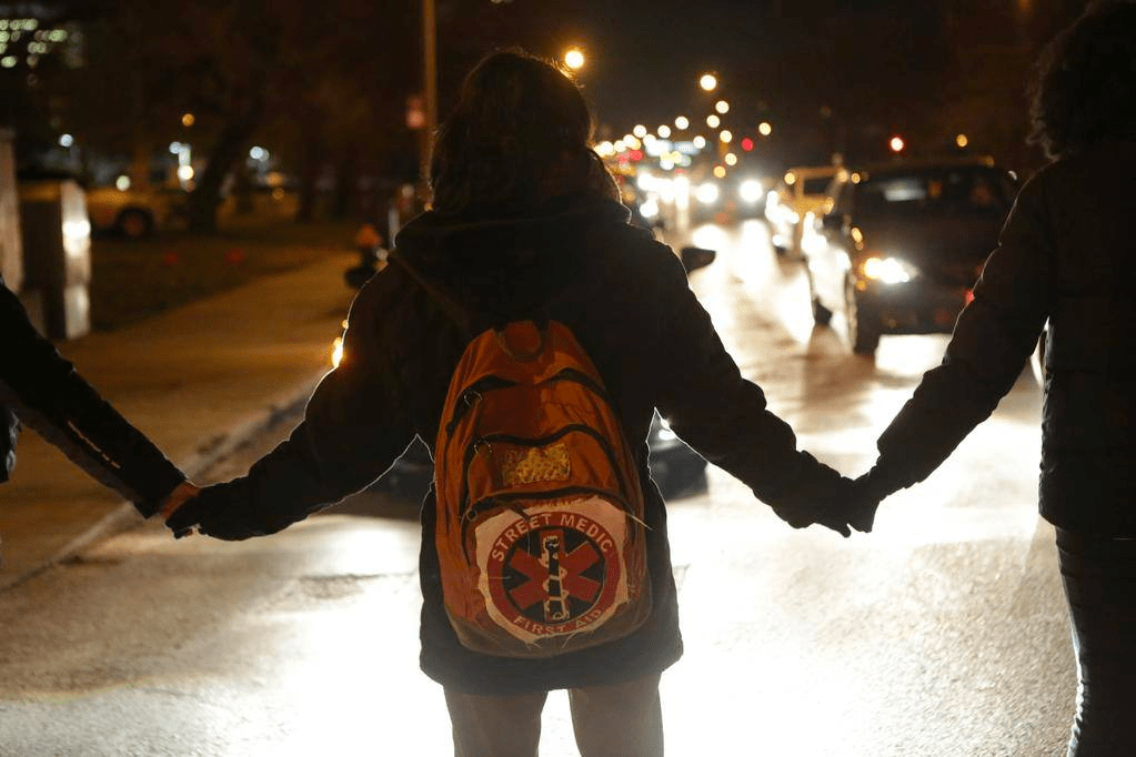 A person with a street medic snake-and-cross patch on their backpack holds hands with other protestors as oncoming traffic approaches. The photo is taken at night.