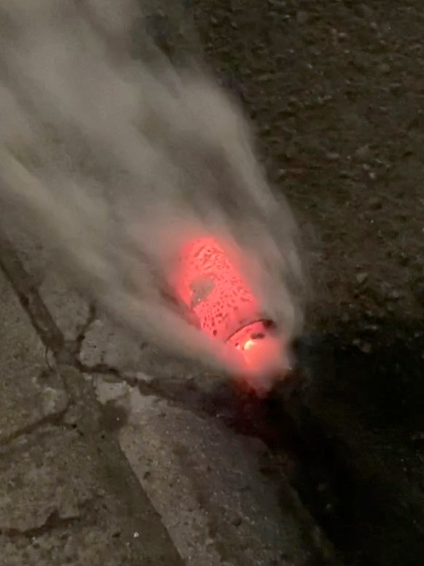 Photo of an active, smoking, glowing red hexachloroethane canister on asphalt.