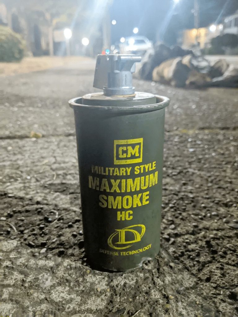 Photo of an inactive, dark-green hexachloroethane canister positioned upright on asphalt. The canister has "CM. Military style maximum smoke HC" printed on it in yellow with a yellow Defense Technology logo below the words.