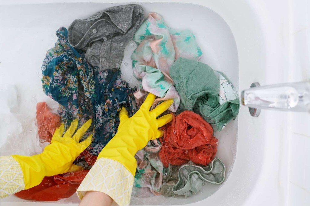 Gloved hands perform a pre-rinse and wash of contaminated clothes of different colors and styles in a bathtub.
